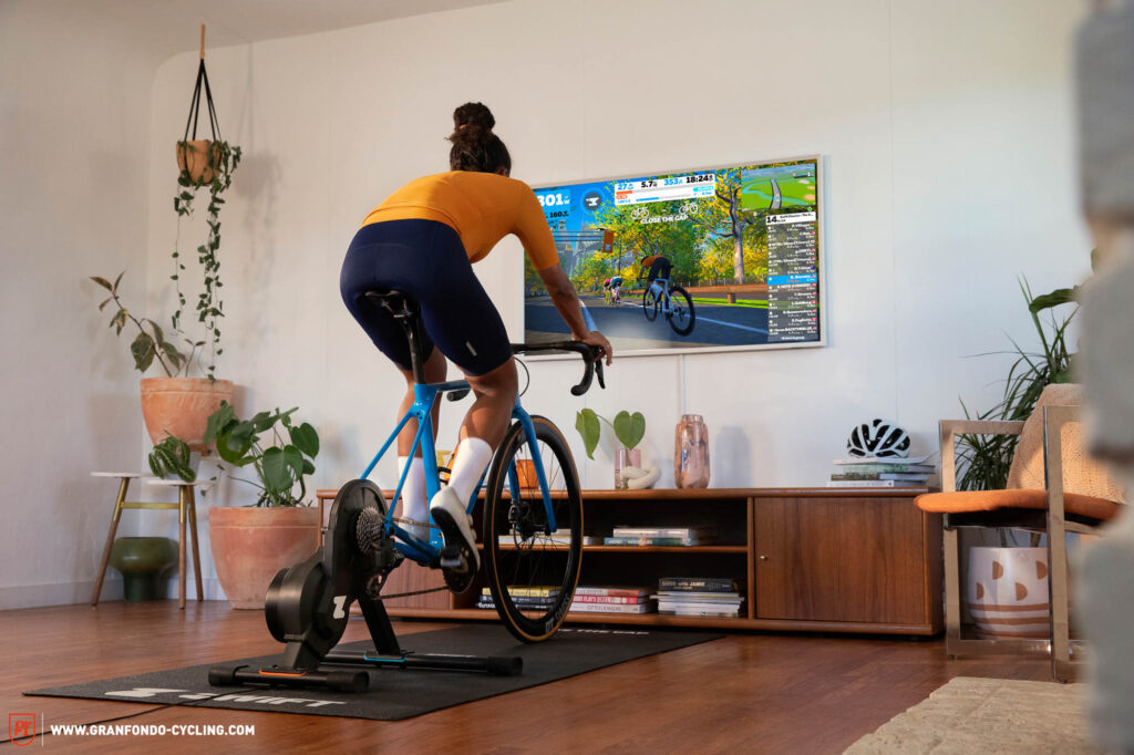 Kvinde cykler indendørs på smart trainer med Zwift på TV-skærm.